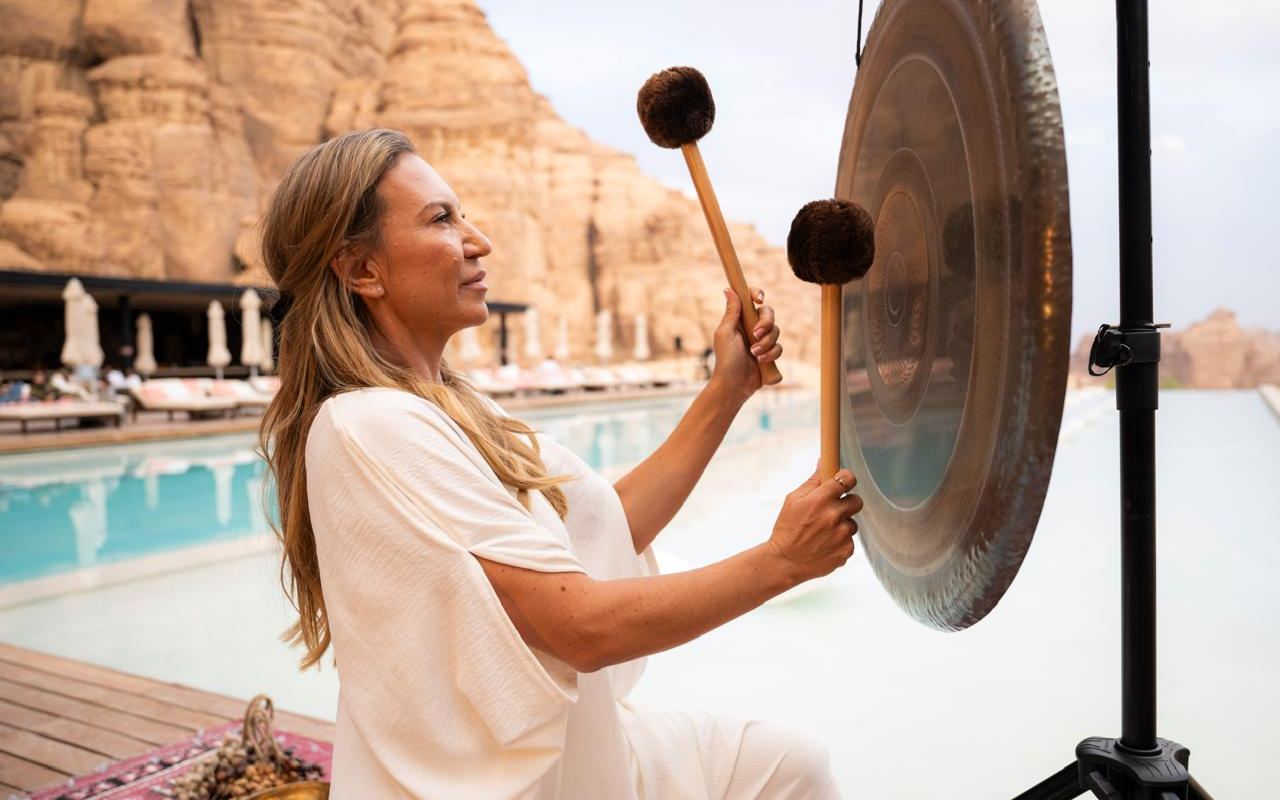 A facilitator sounds a gong beside a still pool at sunset, with the sandstone cliffs of AlUla behind